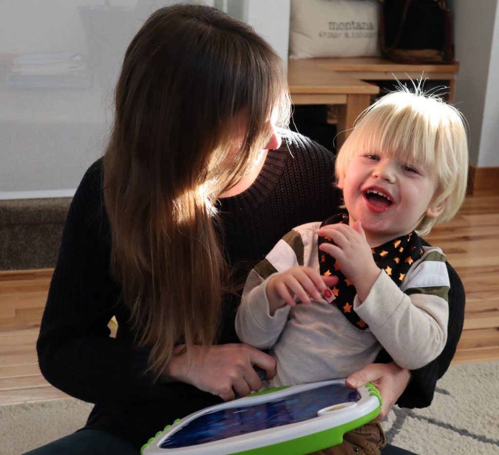 Rebecca, in dark sweater with long hair hanging over her face, holds a laughing Everett, the sunshine coming through a window onto his white-blond hair.