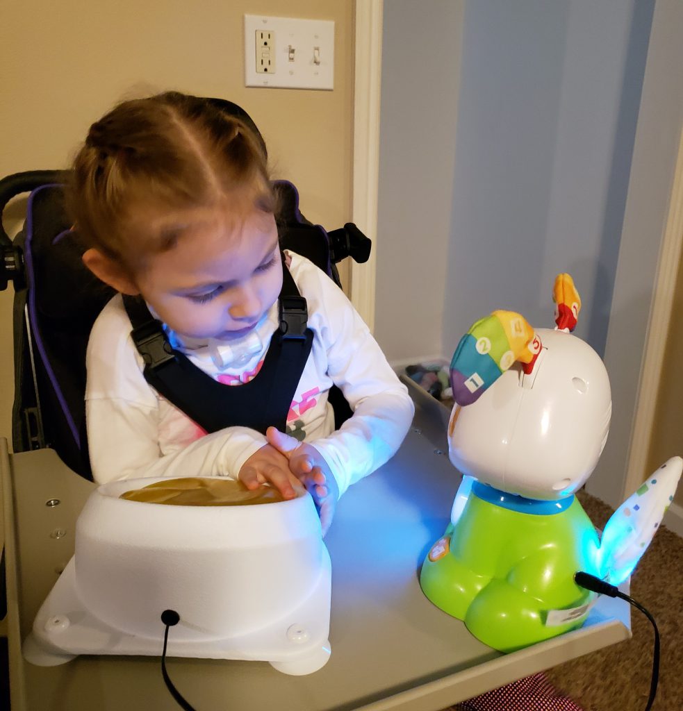 Kinsley sits at tray table and presses a switch to activate the toy she borrowed from MonTECH. Sweet looking little girl with hair in french braids.