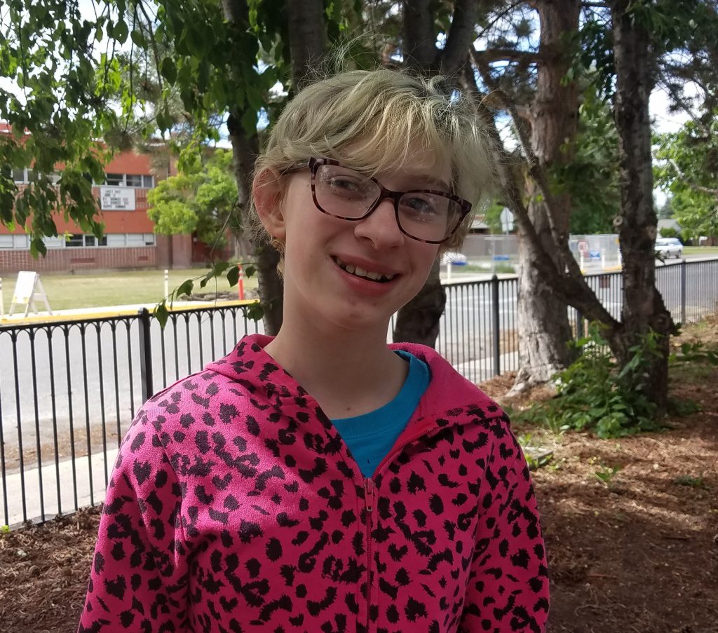 Sarah smiles at camera, wearing hot-pink leopard-spotted hoodie, black frame glasses, and short blond hair.