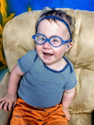 Caleb smiles at the camera, wearing blue plastic glasses, hearing aids on a blue headband, blue striped shirt and orange shorts.