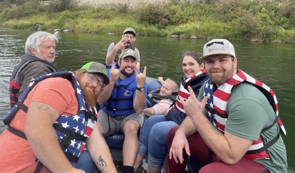 A group of smiling people in llife jackets give rock-on hand gestures while floating down a slow river in a raft. Some have developmental disabilities. Two are wearing life jackets in patriotic stars and stripes.