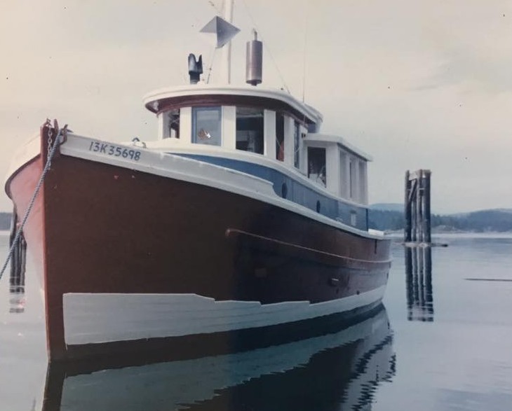 A 50-foot wooden boat, gaily painted rusty red and white, floats on quite waters. A blueish, wooded island is in the distant background.