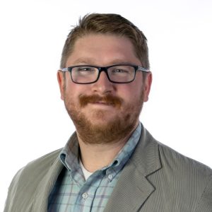 Head shot of Ben Swensen, a young man smiling, wearing suit coat, a beard, and plastic-framed glasses. He has a friendly, good-natured expression.