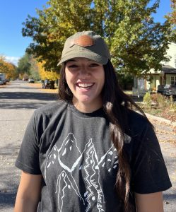 Young woman with long black hair, laughing. She is wearing a ball cap with a leather fish patch on it, and a t-shirt showing a hiking trail on a mountain.