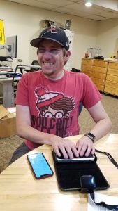 Ed Worrell wears a red Where's Waldo t-shirt and a cap. He's using a refreshable Braille display and laughing. 