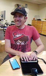 Ed, wearing a Where's Waldo T-shirt and dark cap, laughs while demonstrating a Braille display.