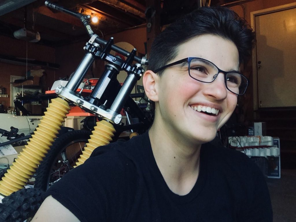 Eleni smiles, wearing black T, black rimmed glasses, short black hair. Customized bicycle in shop behind her.