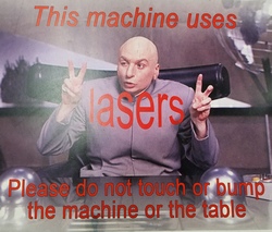 Sign posted on the MakerBot table showing Mike Myers as Dr. Evil, making air quotes as he says "This machine uses lasers. Please do not touch or bump the machine or the table."