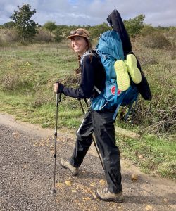 A young woman with braided hair hikes a dirt path. She wears a hat, a big grin, and a big pack on her back with rubber crocs strapped prominently to the side.