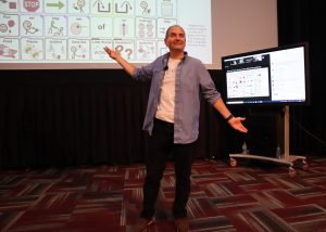 2022 presenter Chris Bugaj smiling and making a tad-dah gesture while standing in front of a huge screen showing communication symbols.