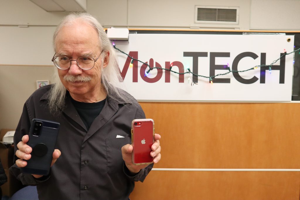 Dave holds up an Android phone and an iPhone. He stands in front of a MonTECH sign festooned with Christmas lights.