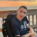 Isaac is sitting in his power chair on a deck. He is smiling. The deck overlooks the ocean and a beautiful glowing sunset is in the background.