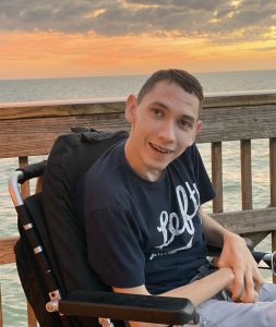 Young man smiles while sitting on deck. The sun sets over the ocean behind him.