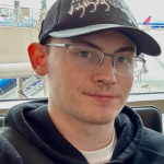 Headshot of a young man in a dark hoodie, wearing glasses and a black billed cap with a cool graphic.