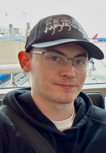 Headshot of a young man in a dark hoodie, wearing glasses and a black billed cap with a cool graphic.