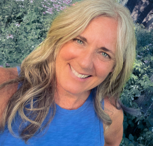 Headshot of a woman smiling, with green shrubbery behind her. She has very blue eyes and long, wavy, blond hair.