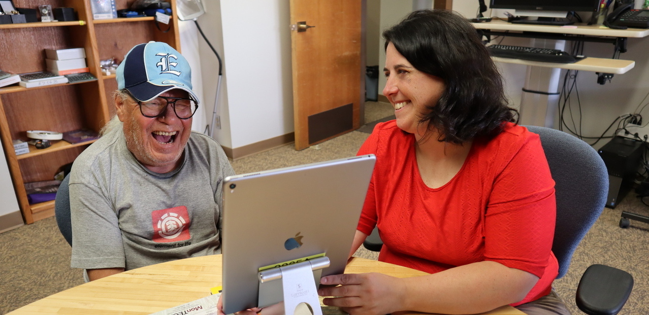 Michelle in red sweater, smiling, works with an iPad and an older man in blue cap, laughing hard.