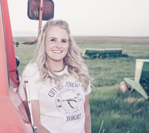 Blonde woman smiles while leaning against a farm truck, a green field with farm machinery behind her.