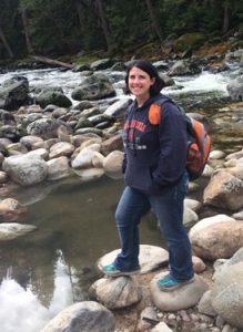 Michelle stands on rocks of creek bed near water, smiling and wearing orange back pack.