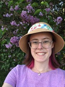 Young woman with red streaks in her hair, glasses, and a straw hat smiles in front of a blooming lilac bush.