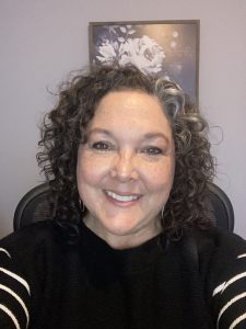Close-up headshot of Paige Buckingham. She has curly dark hair, wears a black sweater, and has a very bright, animated smile.