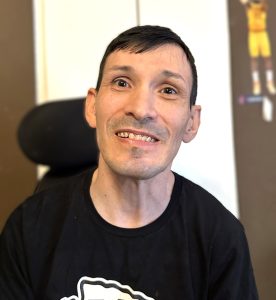 Close-up headshot of a man sitting in a power chair, smiling. He has short dark hair and very bright brown eyes.