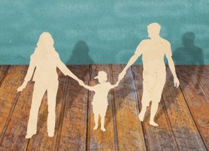 Paper cutouts of two parent figures holding hands with a child. They walk on a wood-slat floor with their shadows on the teal-colored wall behind them.