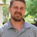 Headshot of Stephen Kneece. He stands relaxed in a polo shirt, dark blond hair, beard, and mustache, with a faint smile for the camera. Greenery and an elegant building are in the background.