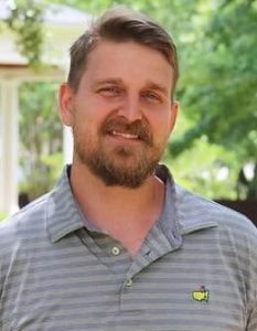 Headshot of Stephen Kneece. He stands relaxed in a polo shirt, dark blond hair, beard, and mustache, with a faint smile for the camera. Greenery and an elegant building are in the background.