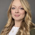 Headshot of Helena. She has very long blond hair in curling waves, big blue eyes, and is smiling with closed lips. She wears a white t-shirt under and jacket and a gold necklace.