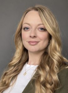 Headshot of Helena. She has very long blond hair in curling waves, big blue eyes, and is smiling with closed lips. She wears a white t-shirt under and jacket and a gold necklace.