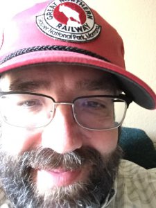 Extreme close-up of Tom with small smile, in red cap, glasses, and full beard and mustache.