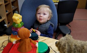 Blond todller pushes button to make jack-in-the-box go, looking at camera saying ooooo.